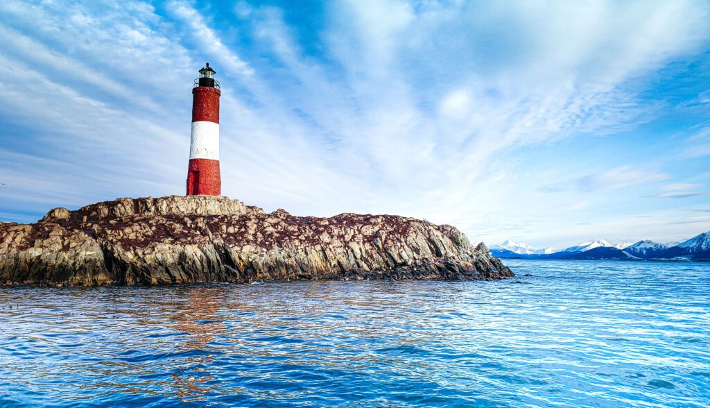 Scenic view of a red and white lighthouse on rocky island with sea and mountains at Ushuaia, Argentina.