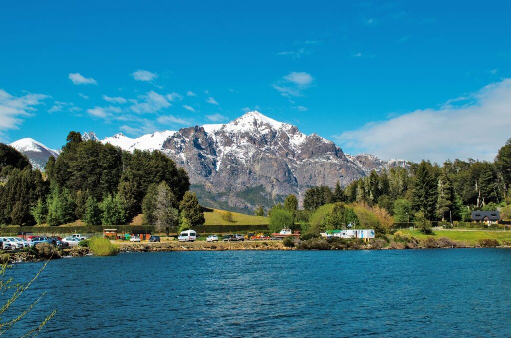 Breathtaking view of snowy mountains and a tranquil lake surrounded by lush greenery under a clear sky.