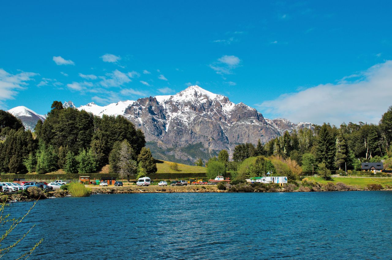 Breathtaking view of snowy mountains and a tranquil lake surrounded by lush greenery under a clear sky.